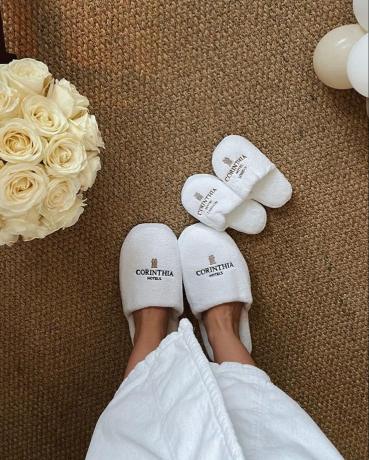 f6844ca3-a33c-4f5b-ad54-627eb11f1919 Cozy white slippers from Corinthia Hotels next to a bouquet of roses on a woven carpet, perfect for relaxation. | Sky Rye Design Cozy white slippers from Corinthia Hotels next to a bouquet of roses on a woven carpet, perfect for relaxation.