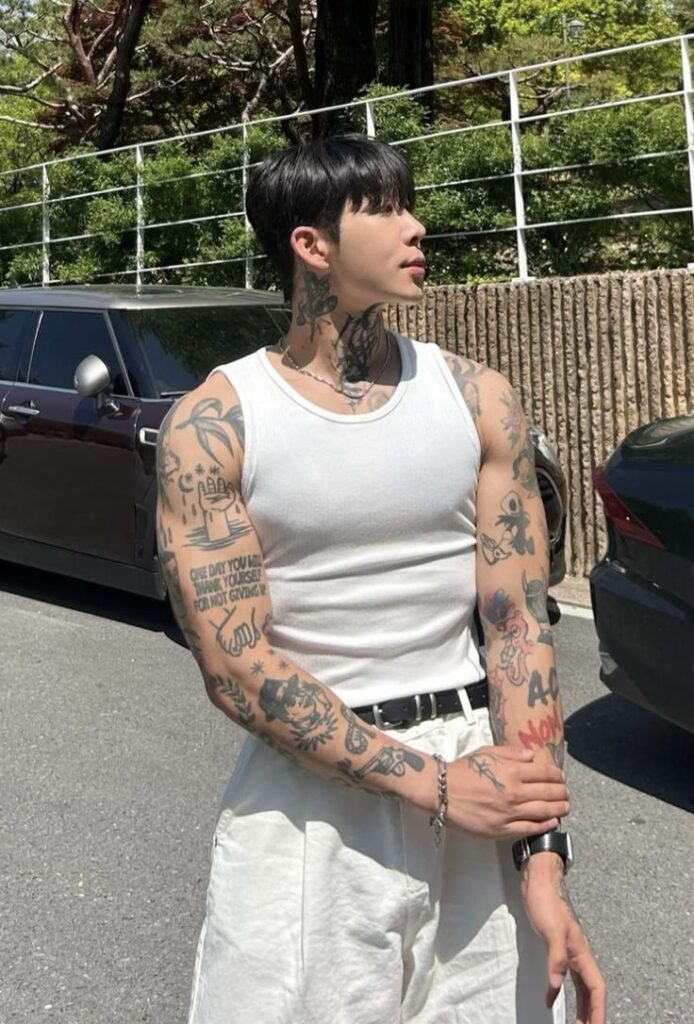 Stylish man with arm tattoos in a white tank top, outdoor setting, showcasing urban fashion.