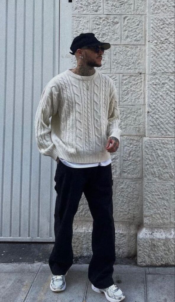 Man in a white cable-knit sweater, black pants, and cap, standing against a textured stone wall on a street sidewalk.
