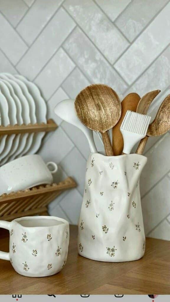 Ceramic mug and utensil holder with wooden kitchen tools on a counter, against a tiled backsplash.