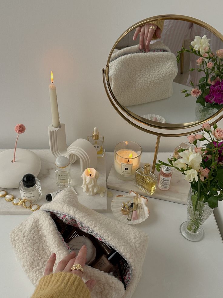 Vanity setup with flowers, candles, skincare products, and an open toiletry bag reflected in a round mirror.