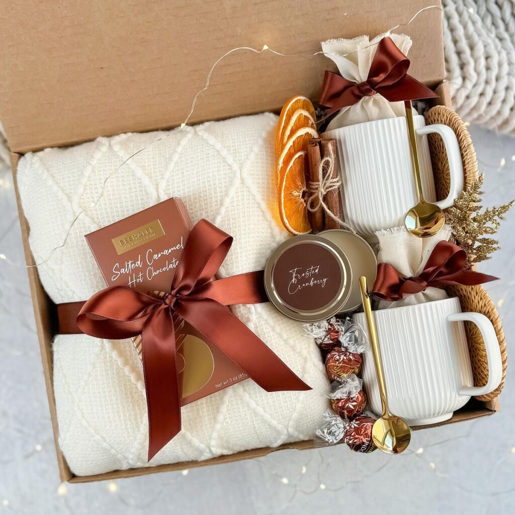 Cozy gift box with mugs, hot chocolate, candles, and treats, wrapped in festive ribbons, perfect for holidays.