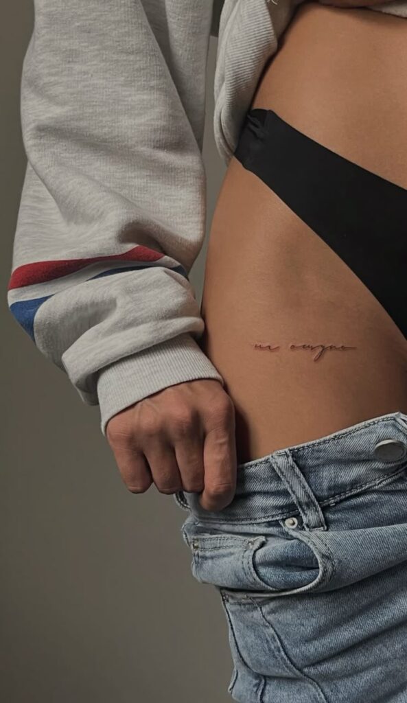 feaf57d94afb2400d2d5b18b545373f2 Close-up of a side hip tattoo with text, person wearing jeans and a sweatshirt, showcasing minimalist body art. | Sky Rye Design Close-up of a side hip tattoo with text, person wearing jeans and a sweatshirt, showcasing minimalist body art.