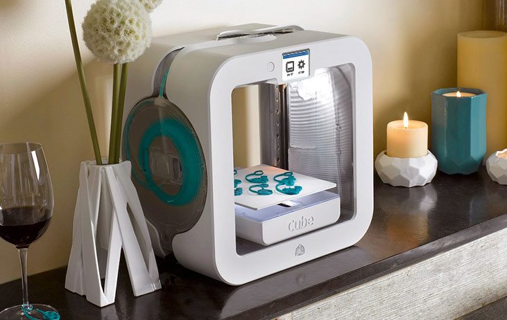 Compact 3D printer on a shelf with candles, a vase, and a glass of red wine, printing turquoise objects.