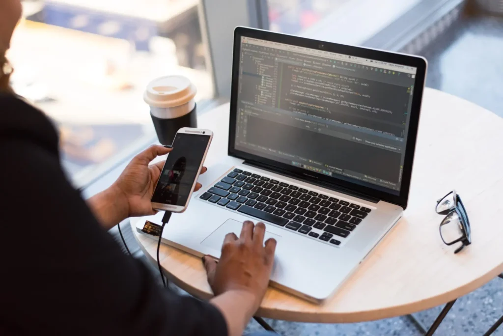 Person coding on laptop while holding a smartphone, coffee and glasses on table, suggesting a work environment.