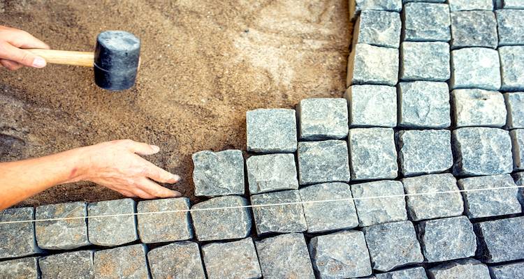 fitting-brick-stone-steps-cost-4-c2m800 Person laying cobblestones with a mallet on a sandy surface, aligning with string for precise placement. | Sky Rye Design Person laying cobblestones with a mallet on a sandy surface, aligning with string for precise placement.