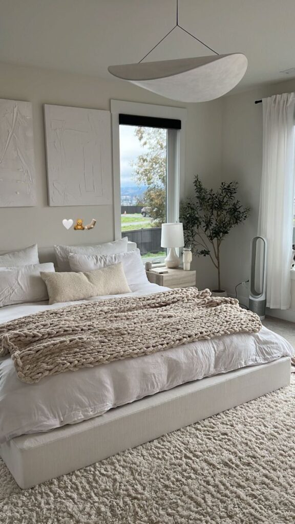 Modern bedroom with cozy bedding, large window view, white decor, and a knitted blanket on a plush rug.