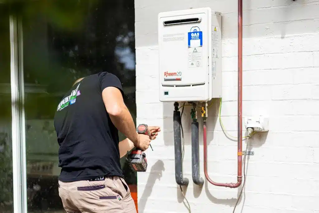 Technician installing a Rheem gas water heater on an exterior brick wall.