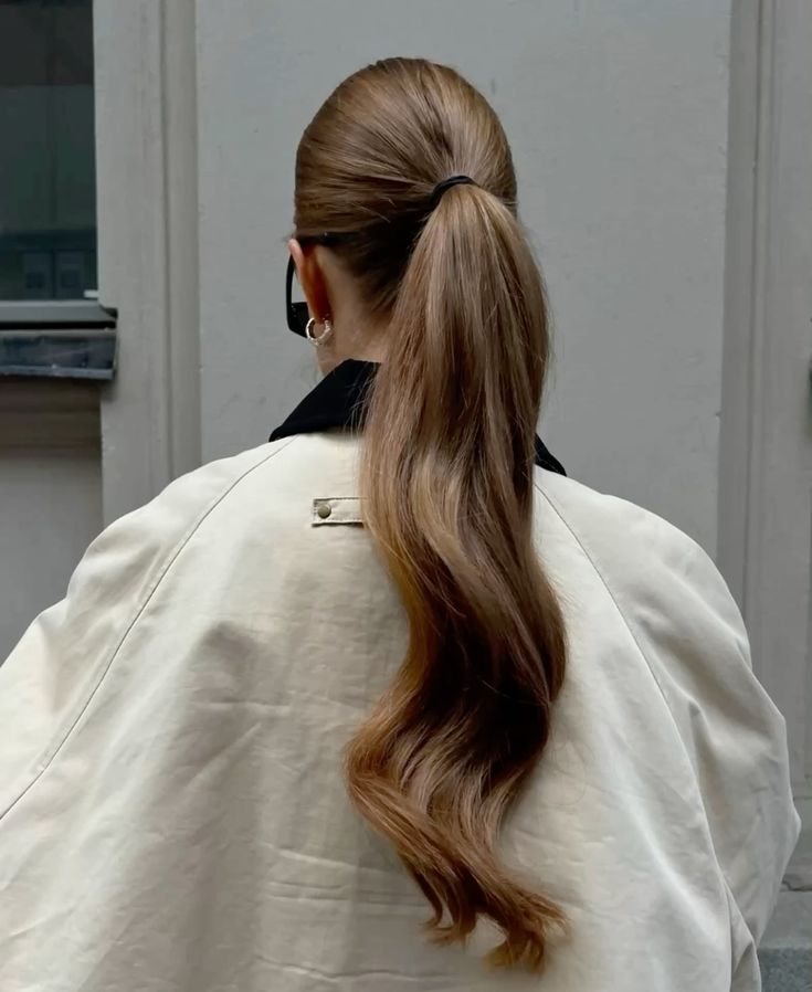 Woman with long, wavy brown hair in a ponytail, wearing a white coat, viewed from behind.