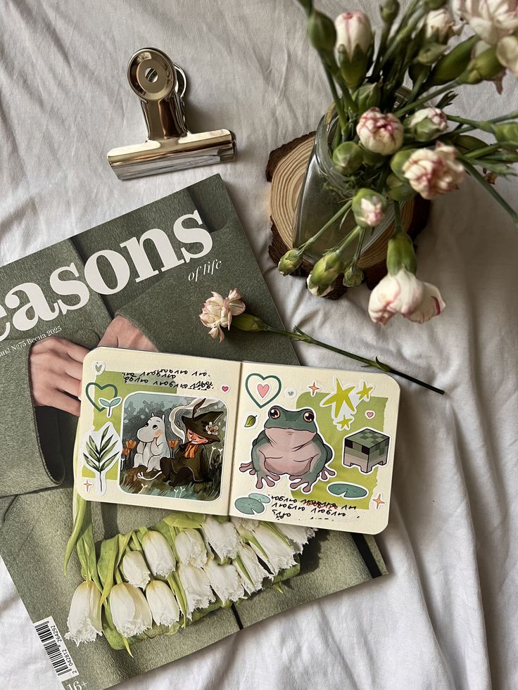 Open journal with illustrations of a frog and character, surrounded by flowers and a magazine on a white cloth.