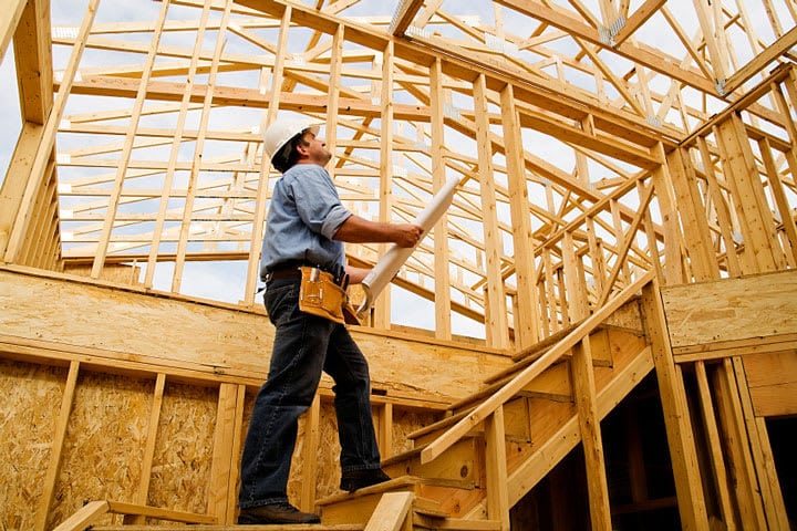 home-builder-720_f7wmln Construction worker examining house frame with plans, wearing a hard hat and tool belt, on wooden staircase. | Sky Rye Design Construction worker examining house frame with plans, wearing a hard hat and tool belt, on wooden staircase.
