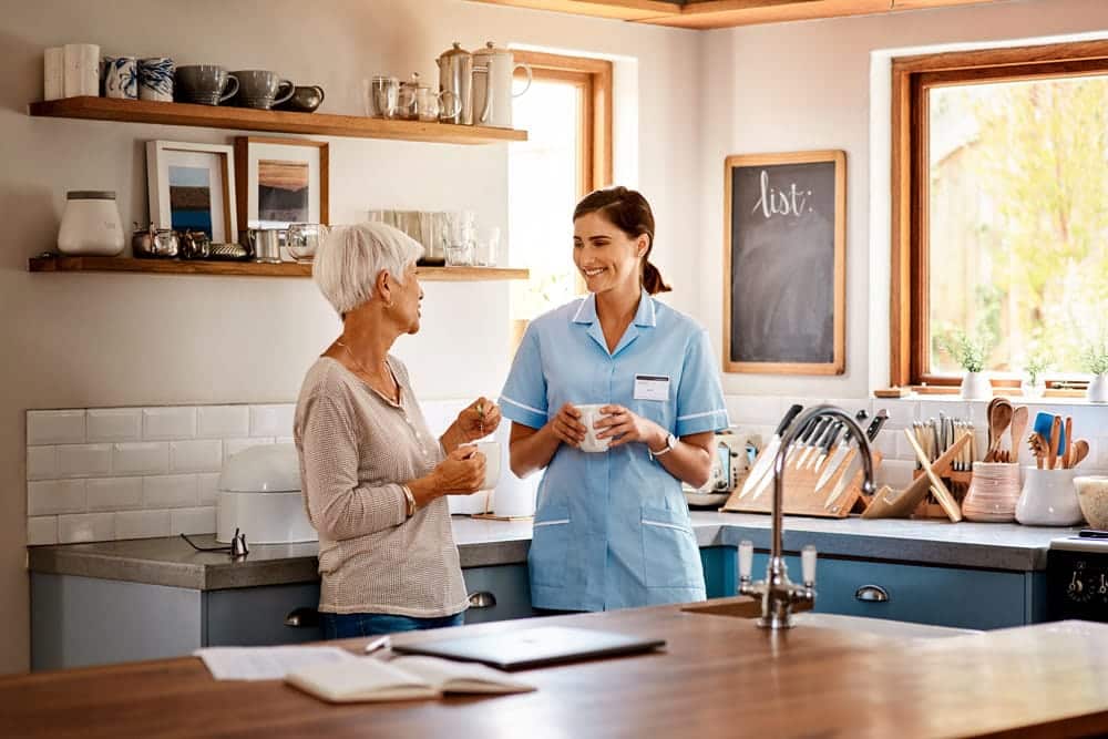 Senior woman and caretaker enjoy a conversation in a bright kitchen, fostering a friendly and supportive environment.