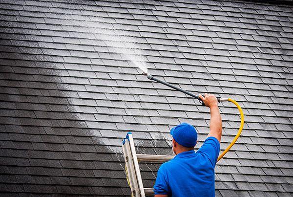 Worker in blue cleaning roof shingles with a pressure washer, removing dirt and grime.