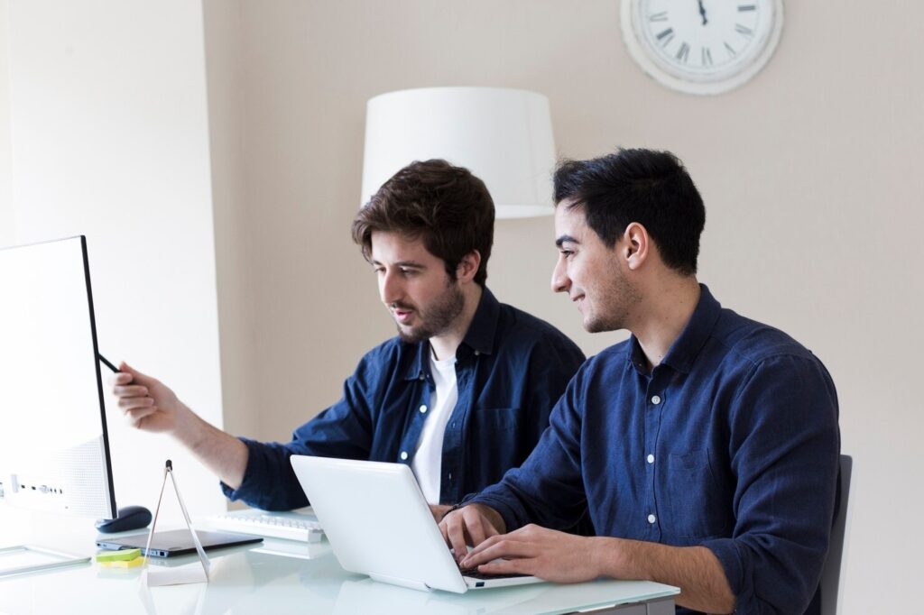 Two men collaborating at a desk with a laptop and computer, focused on work in a modern office setting.
