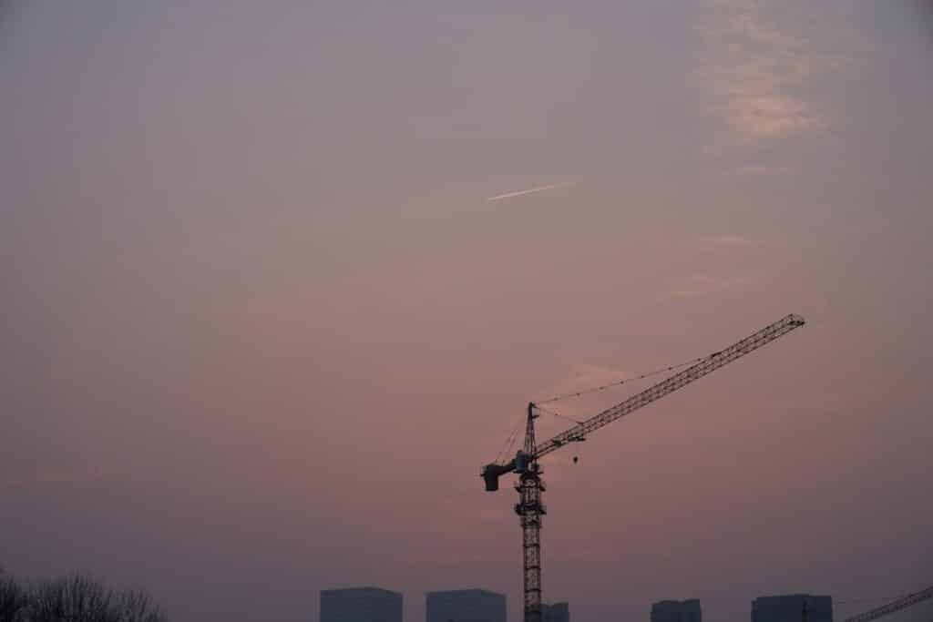 Construction crane silhouetted against a pink sunset sky with city skyline in the background.