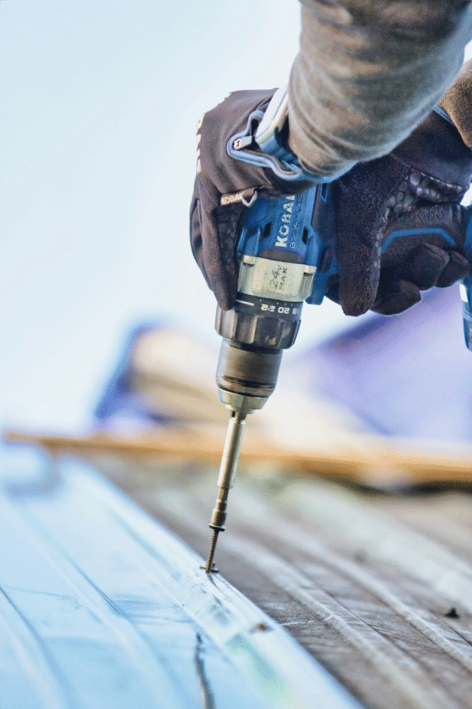Worker using a power drill to secure metal roofing panel with screws, close-up of tool and gloved hands.