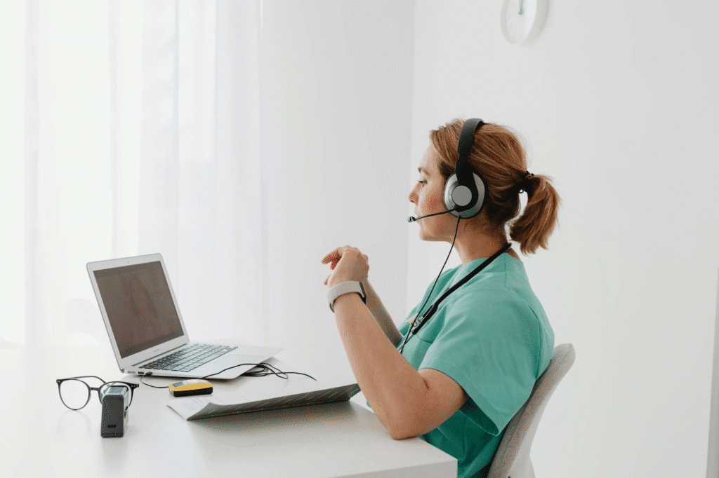Healthcare professional videoconferencing on laptop, wearing headset, in a well-lit room with medical equipment.