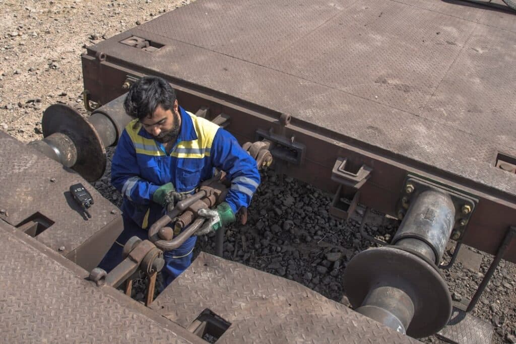Railway worker in blue overalls adjusting coupler between two train cars on a sunny day.