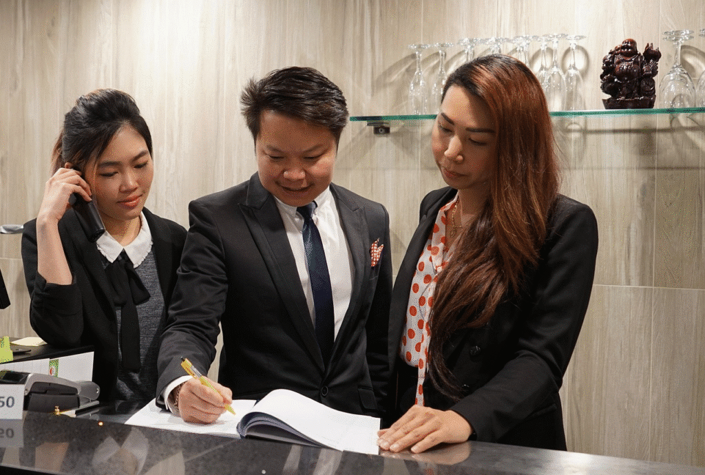 Business team reviewing documents at a modern reception desk, with woman on phone and glasses in the background.