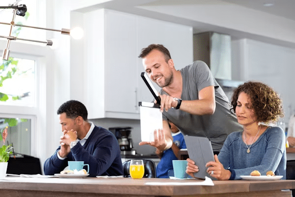 image Team collaboration in a modern kitchen setting, with a man holding a tablet and others engaged in discussion and research. | Sky Rye Design Team collaboration in a modern kitchen setting, with a man holding a tablet and others engaged in discussion and research.