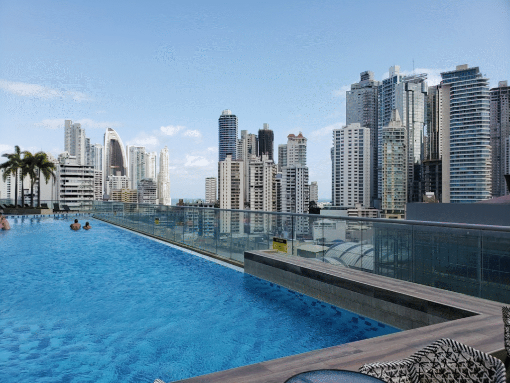 Rooftop infinity pool with stunning city skyline view, high-rise buildings, and clear blue sky in the background.