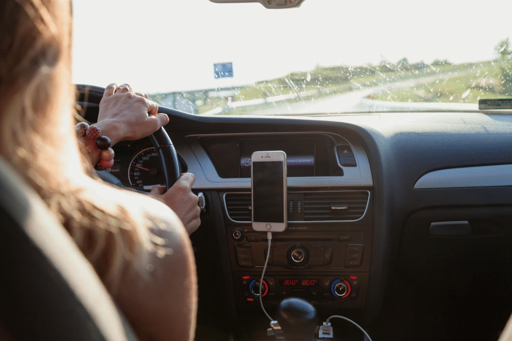 Person driving a car with GPS navigation on phone mounted to dashboard. Sunlit highway view through windshield.