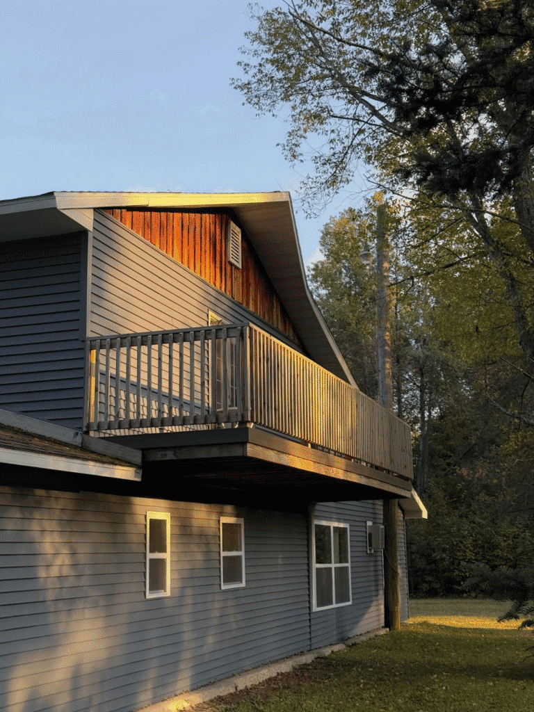 Elevated wooden balcony on a modern grey house exterior during golden hour, surrounded by lush green trees.