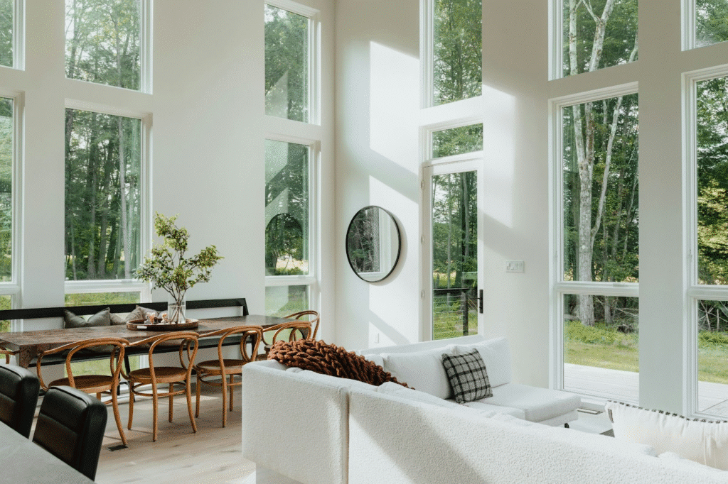 Bright modern living and dining area with large windows, white sofa, wooden table, and lush green outdoor view.