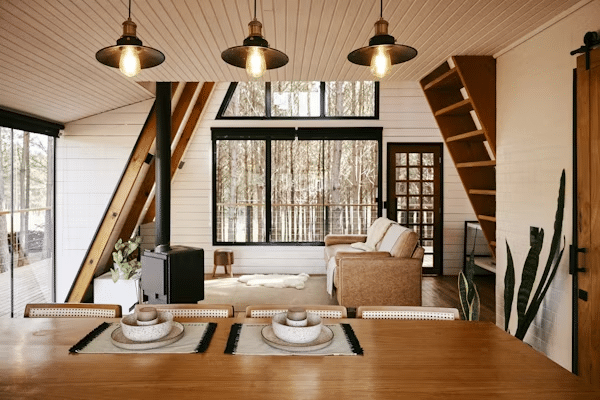 A-frame cabin interior with cozy living area, wooden accents, and large windows offering forest views.