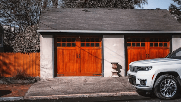 White SUV parked in front of a house with two wooden garage doors and a white planter on the driveway.