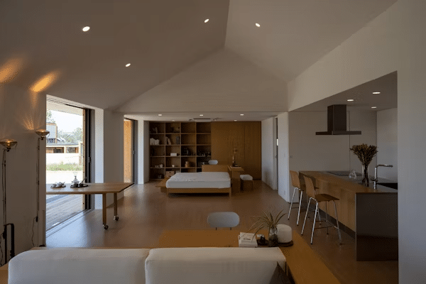 Modern open-plan studio apartment with wooden interiors, featuring a bedroom, kitchen, and natural light from large windows.