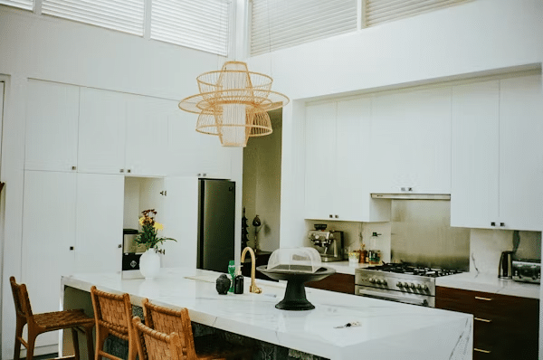 Modern kitchen with white cabinets, marble island, wicker chairs, and a stylish pendant light above.