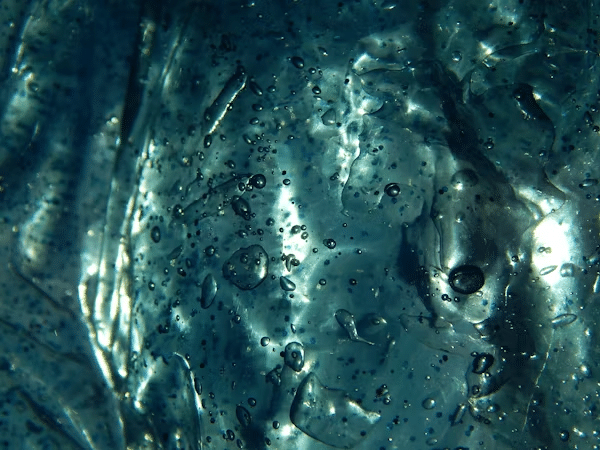 Close-up of clear blue ice texture with air bubbles and light reflections, creating a dynamic, abstract pattern.