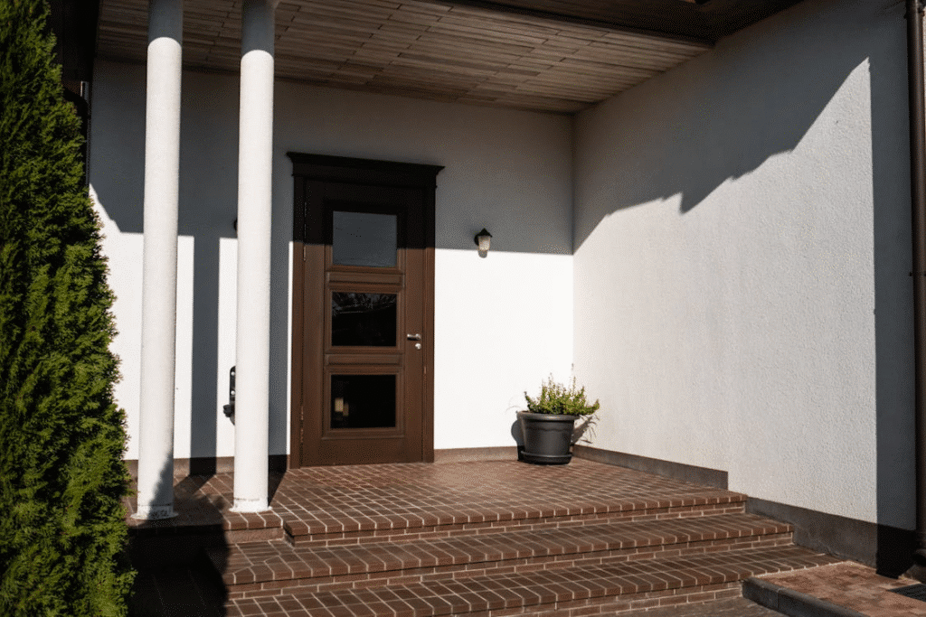 image Modern house entrance with wooden door, brick steps, and potted plant, under sunlight. | Sky Rye Design Modern house entrance with wooden door, brick steps, and potted plant, under sunlight.