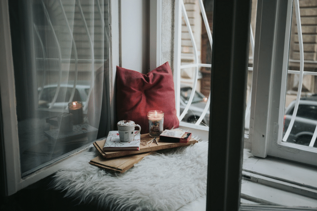 Cozy window nook with candle, books, and coffee on a fluffy surface, perfect for relaxing and reading.