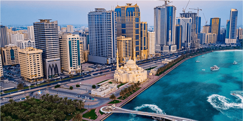 Aerial view of Sharjah's skyline with skyscrapers, waterfront, and a mosque surrounded by lush greenery at sunset.