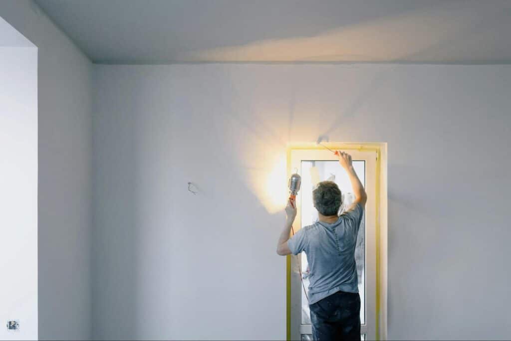 Man painting around a window with a roller, holding a light, in a bright room.