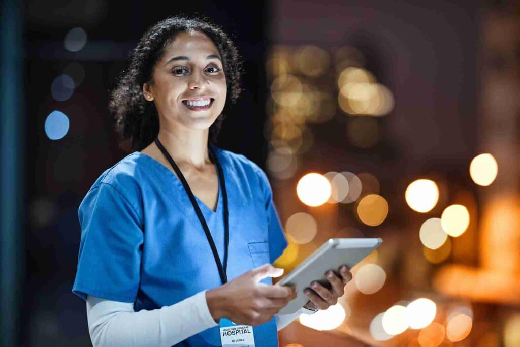 image50f6c631f35684a029497e1546e914aa69f88838 Smiling healthcare worker in blue scrubs holding tablet, city lights background. | Sky Rye Design Smiling healthcare worker in blue scrubs holding tablet, city lights background.