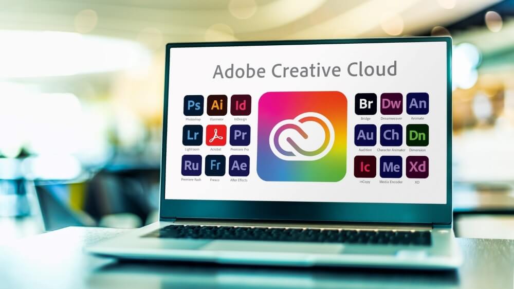 Laptop displaying Adobe Creative Cloud icons, including Photoshop, Illustrator, InDesign, and more on a blurred background.
