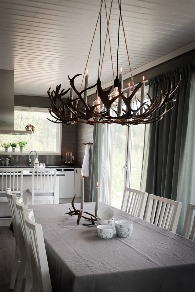 Rustic dining room with antler chandelier, wooden chairs, and gray tablecloth next to large windows and modern kitchen.