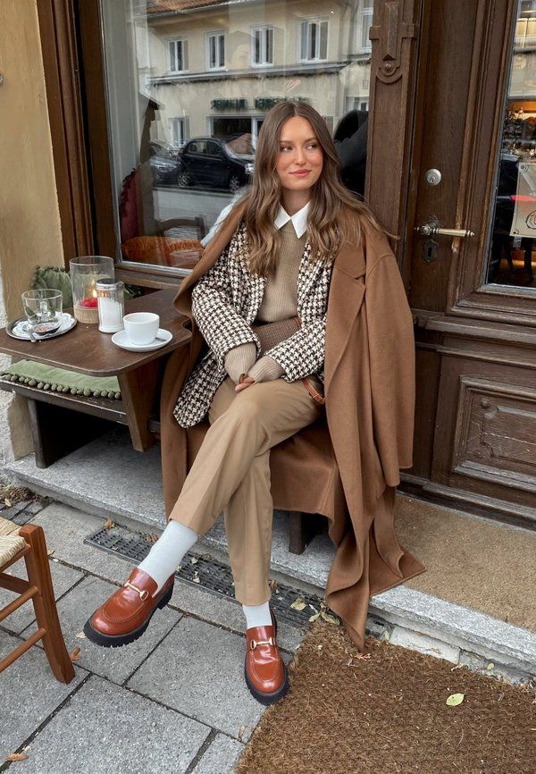 Woman in stylish brown outfit sitting at a café outdoor table with coffee, houndstooth jacket, and loafers.