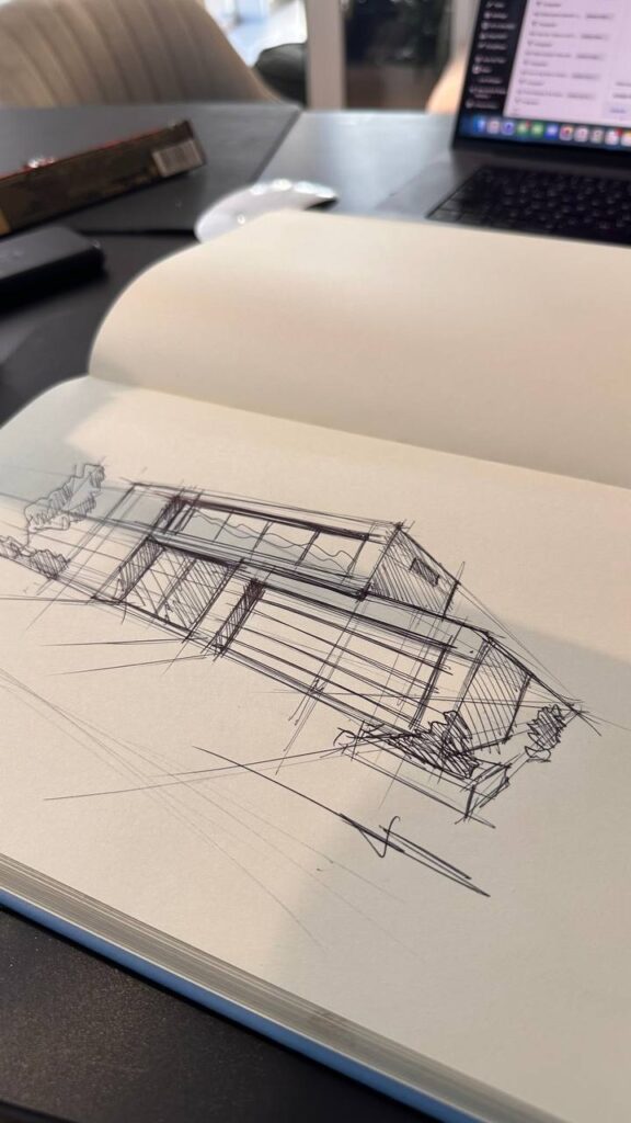 photo_2026-05-27_17-15-32 Architectural pen sketch of a modern house in an open sketchbook on a desk workspace with laptop and mouse | Sky Rye Design Architectural pen sketch of a modern house in an open sketchbook on a desk workspace with laptop and mouse