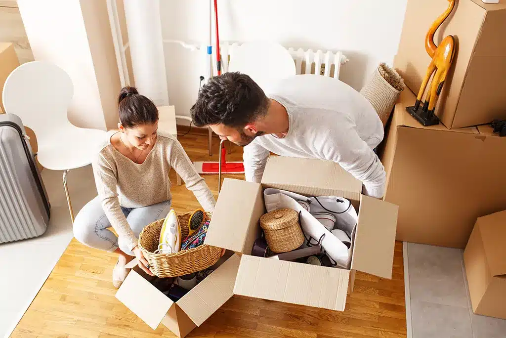 moving-hacks Couple packing household items into boxes for a move, surrounded by packed cartons and luggage in a bright room. | Sky Rye Design Couple packing household items into boxes for a move, surrounded by packed cartons and luggage in a bright room.