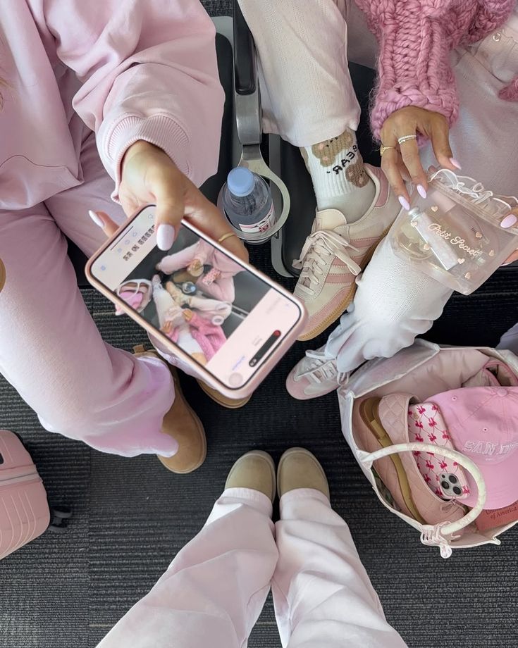 People in pink outfits relax at an airport, one holds a phone, capturing the moment, surrounded by bags and shoes.