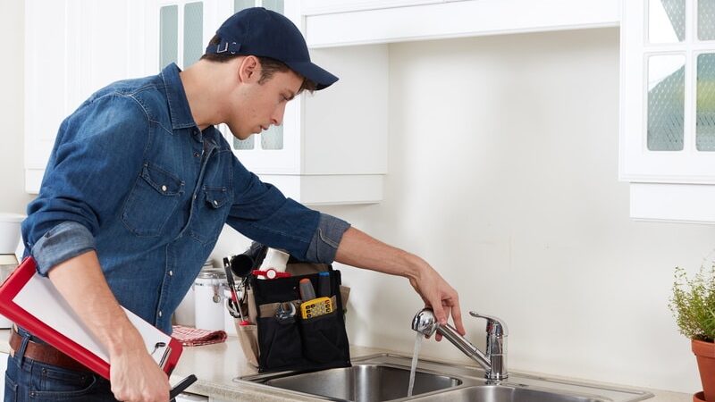 Plumber inspecting kitchen sink faucet in modern kitchen, holding clipboard and tools.