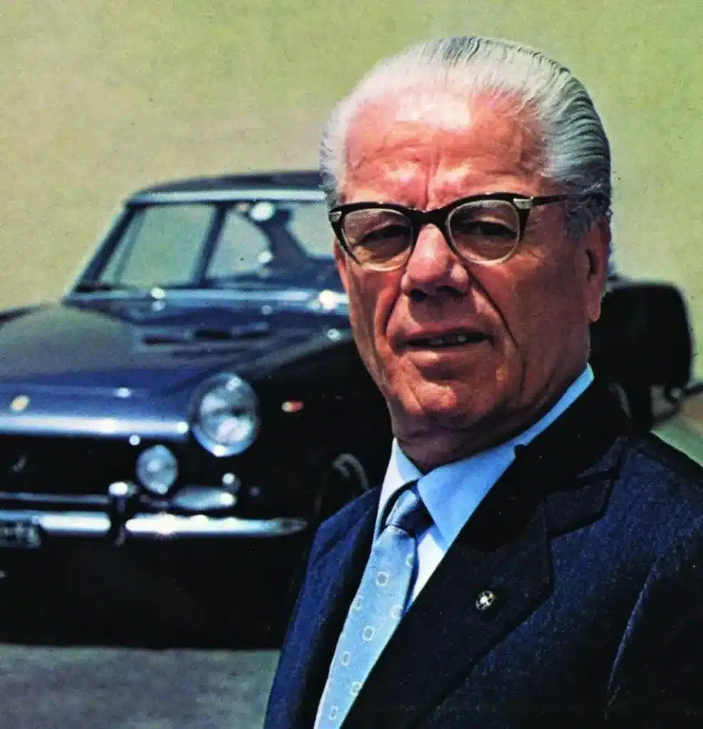 Elderly man in suit standing in front of a classic black sports car with shiny paint and vintage design.