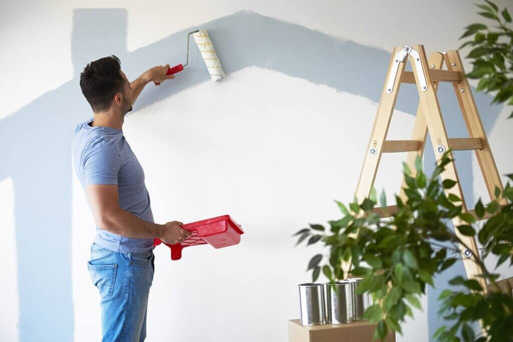 Man painting a wall with a roller, standing near a ladder and paint cans, with green plant in foreground.