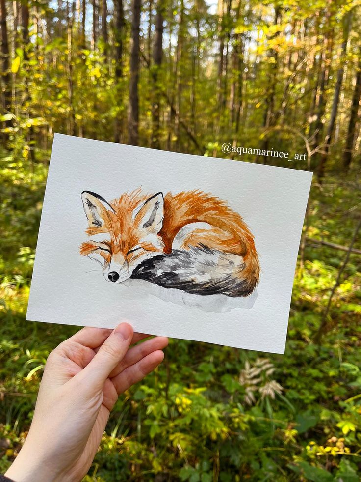 A hand holds a watercolor painting of a sleeping fox in a forest, showcasing nature and art together.