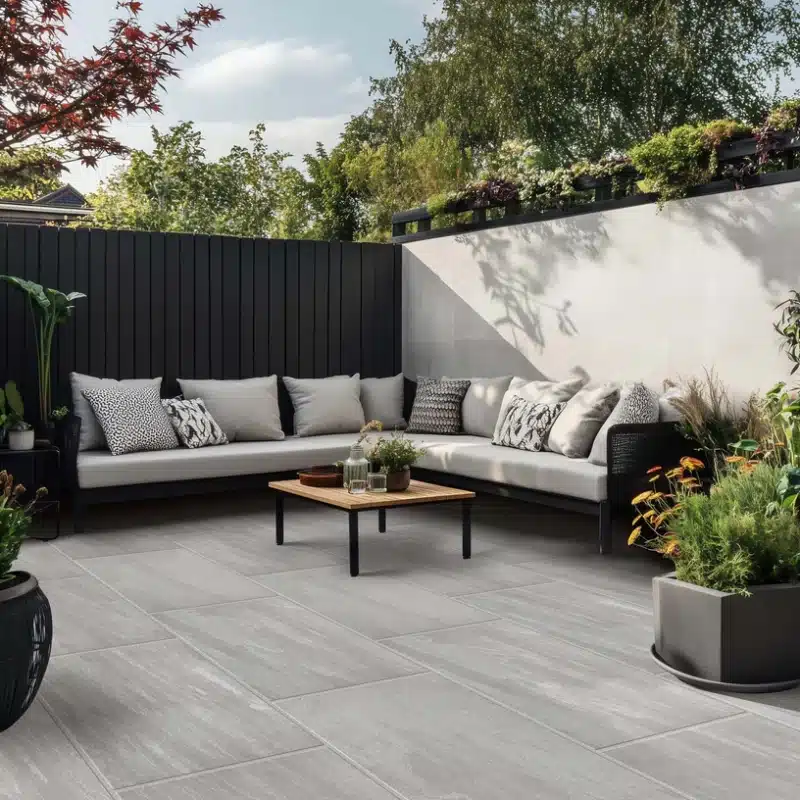 Modern patio with gray tiles, cozy corner sofa, cushions, and lush greenery, creating a serene outdoor space.