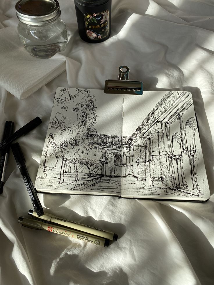Open sketchbook on white fabric, featuring an architectural drawing. Nearby, pens, a jar, and a candle add ambiance.